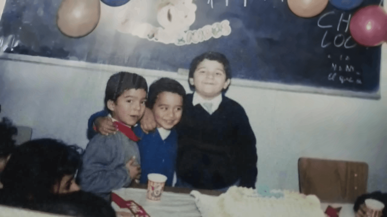 Tres niños sonrientes, abrazados, posan frente al pizarrón decorado con globos durante un festejo en un aula del Instituto